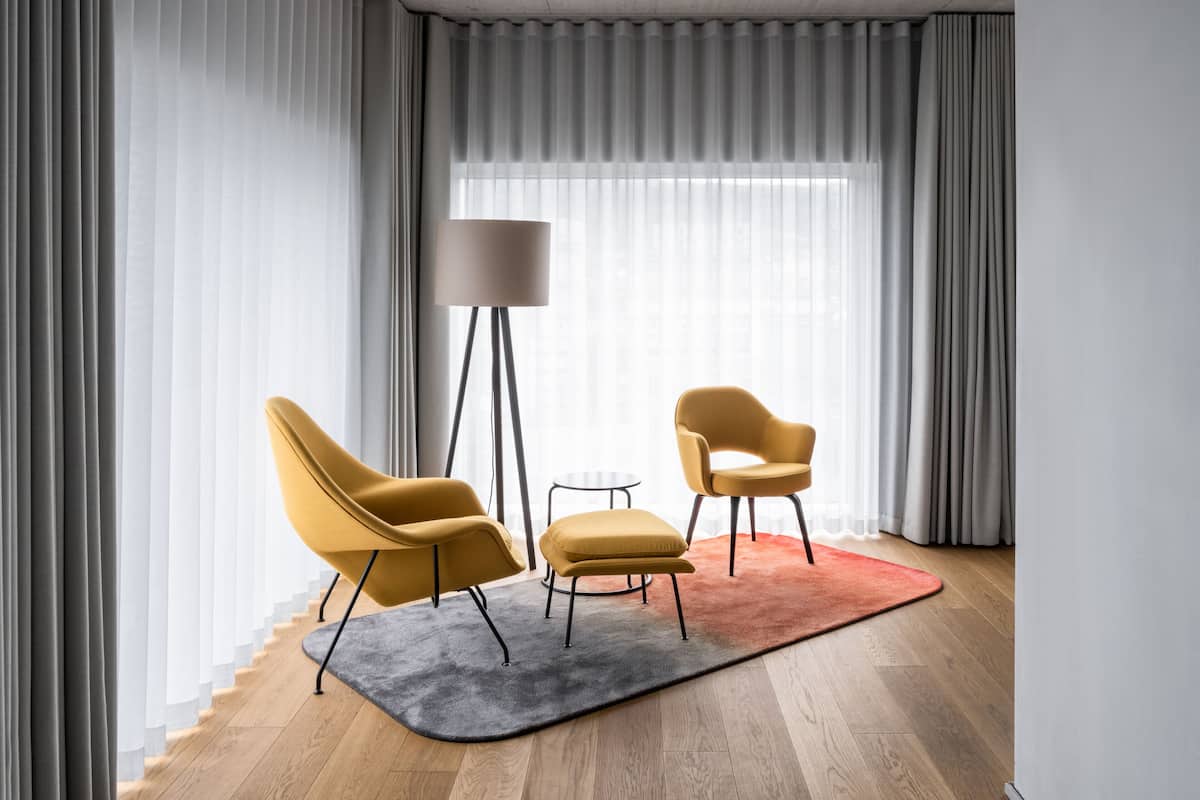 Un salon moderne avec une grande fenêtre, deux fauteuils, un petit pouf et une lampe sur pied.