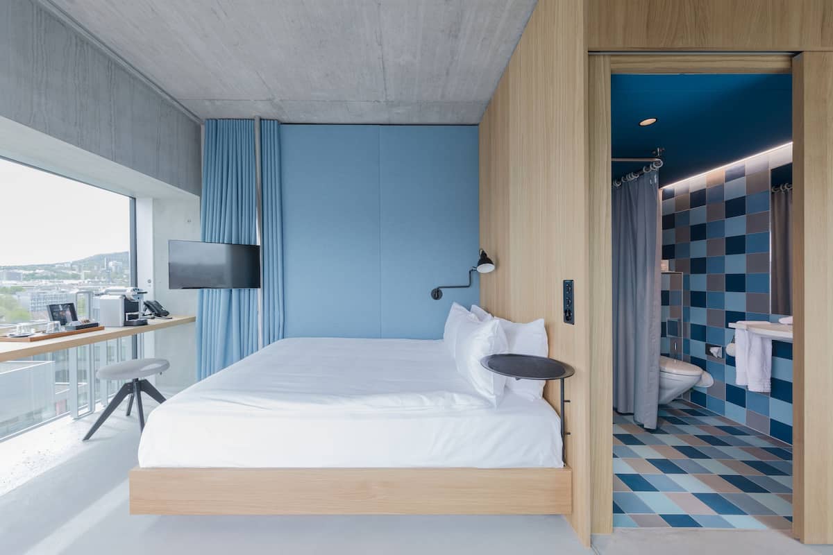 Une chambre d’hôtel moderne dotée d’un grand lit, d’un bureau avec un téléviseur à écran plat, d’une vue sur la ville et d’une salle de bain au sol à carreaux.