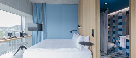 A modern hotel room with a large bed, a desk with a flat-screen TV, a view of the city, and a bathroom with a checkered floor.