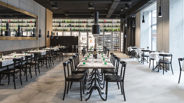 Un vaste espace dédié à la salle à manger, avec de longues tables disposées pour accueillir de nombreux convives, entouré d’étagères remplies de bouteilles et de verres.