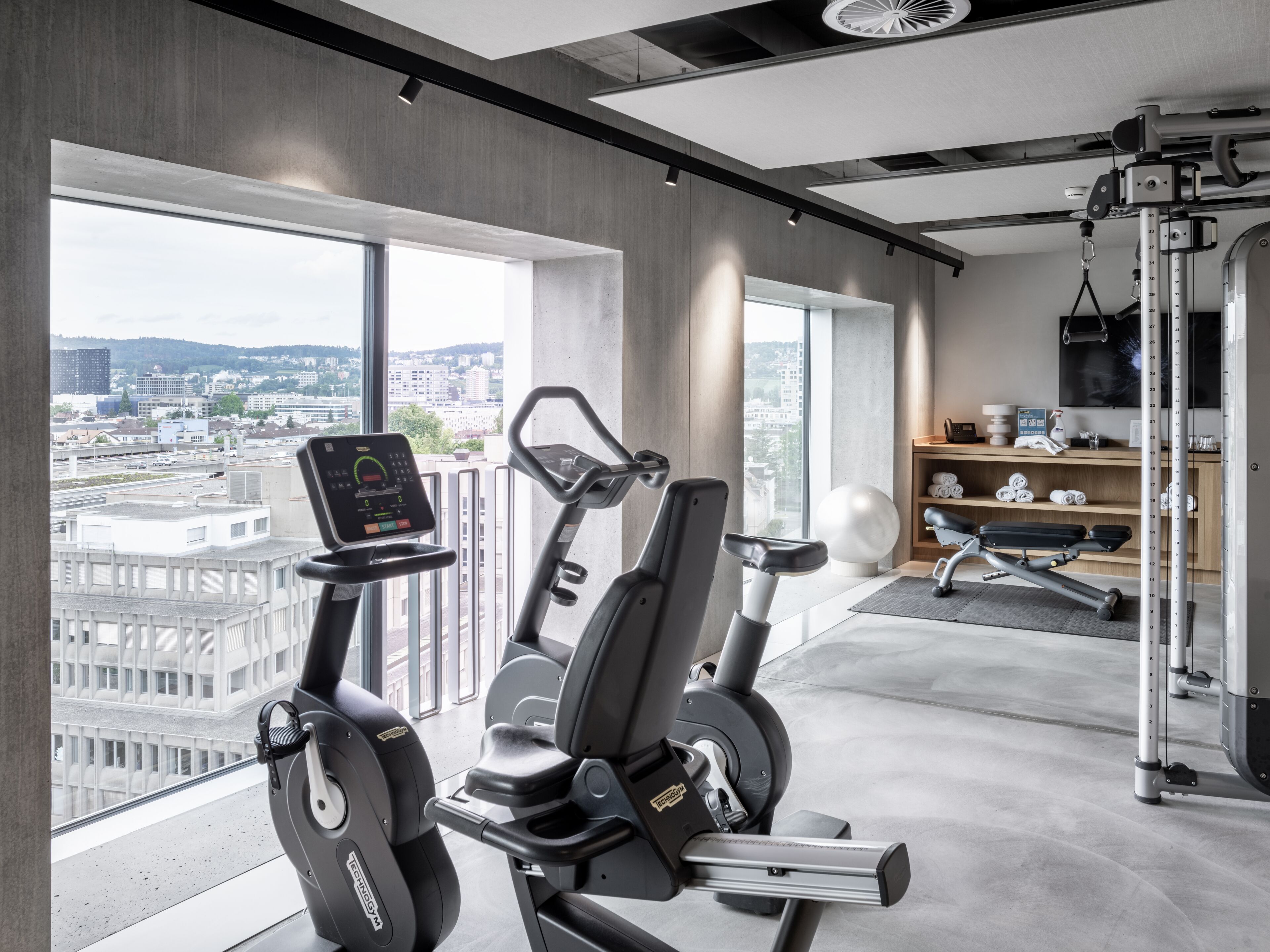 A modern gym with cardio equipment, a weight bench, and a view of the city.