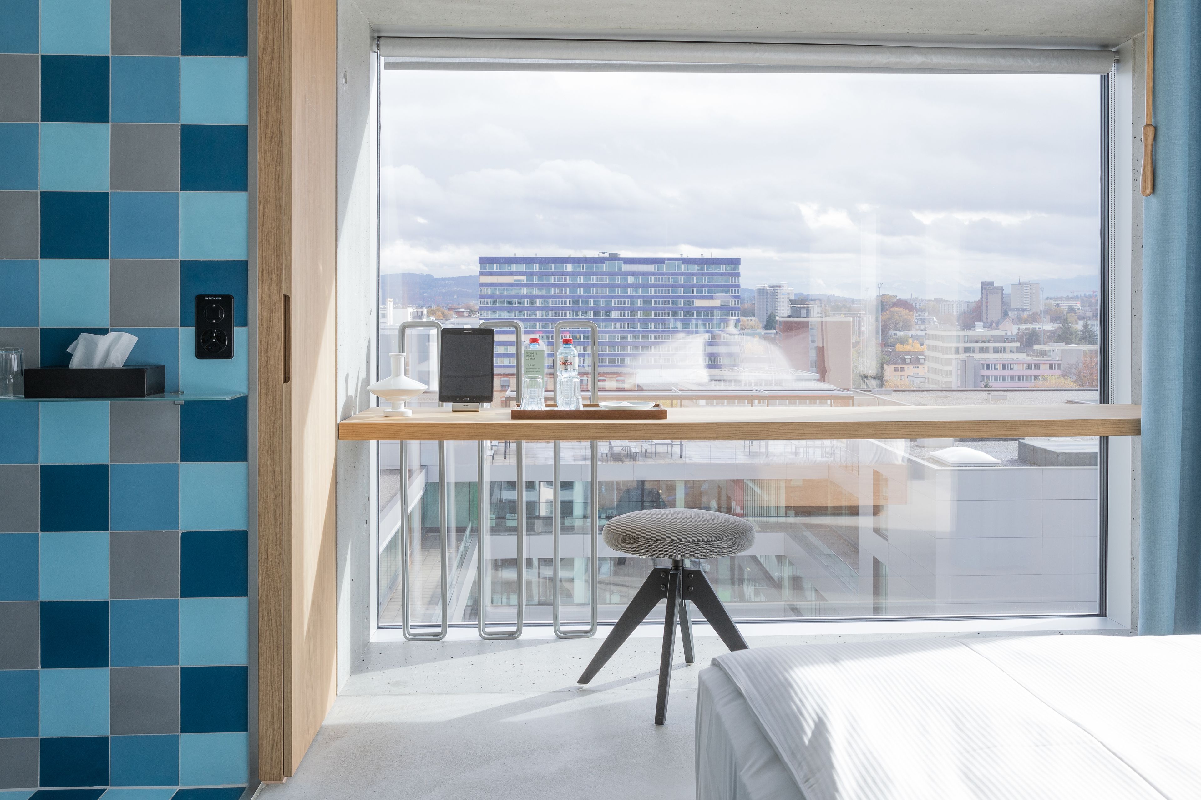 Een moderne hotelkamer met een groot raam, een bureau met een laptop, een waterfles en een klein krukje.