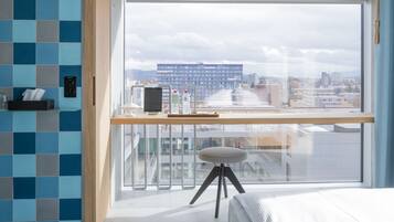 Een moderne hotelkamer met een groot raam, een bureau met een laptop, een waterfles en een klein krukje.