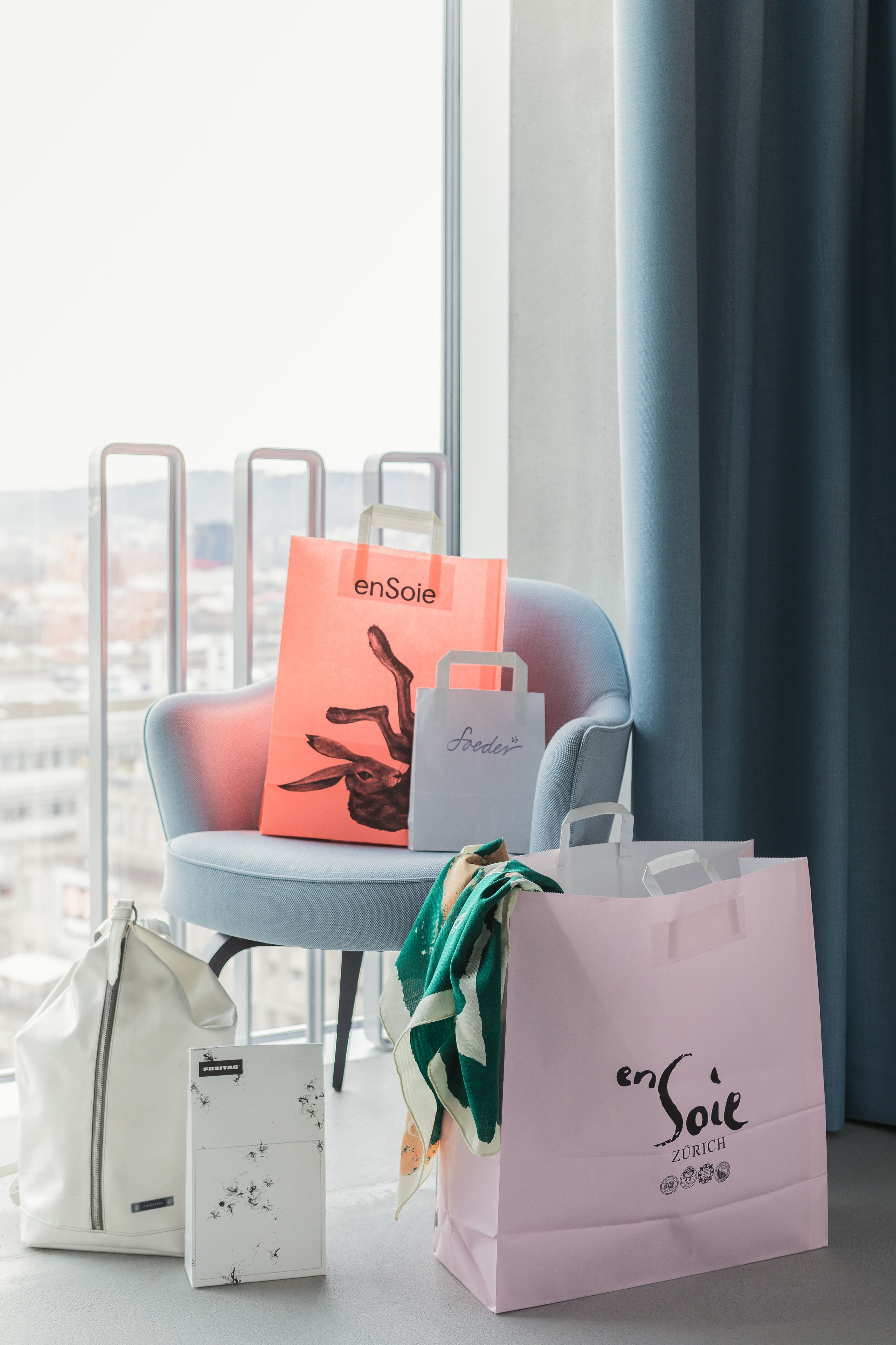 A chair with shopping bags and a box in front of a window with a city view.