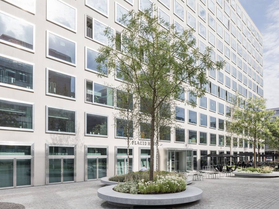 A modern building with a glass facade, a central courtyard with trees and seating, and a sign reading "PLACID HOTEL".