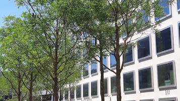 Un edificio moderno con techo plano y ventanas amplias. Hay árboles frente al edificio, y asientos exteriores con sombrillas.