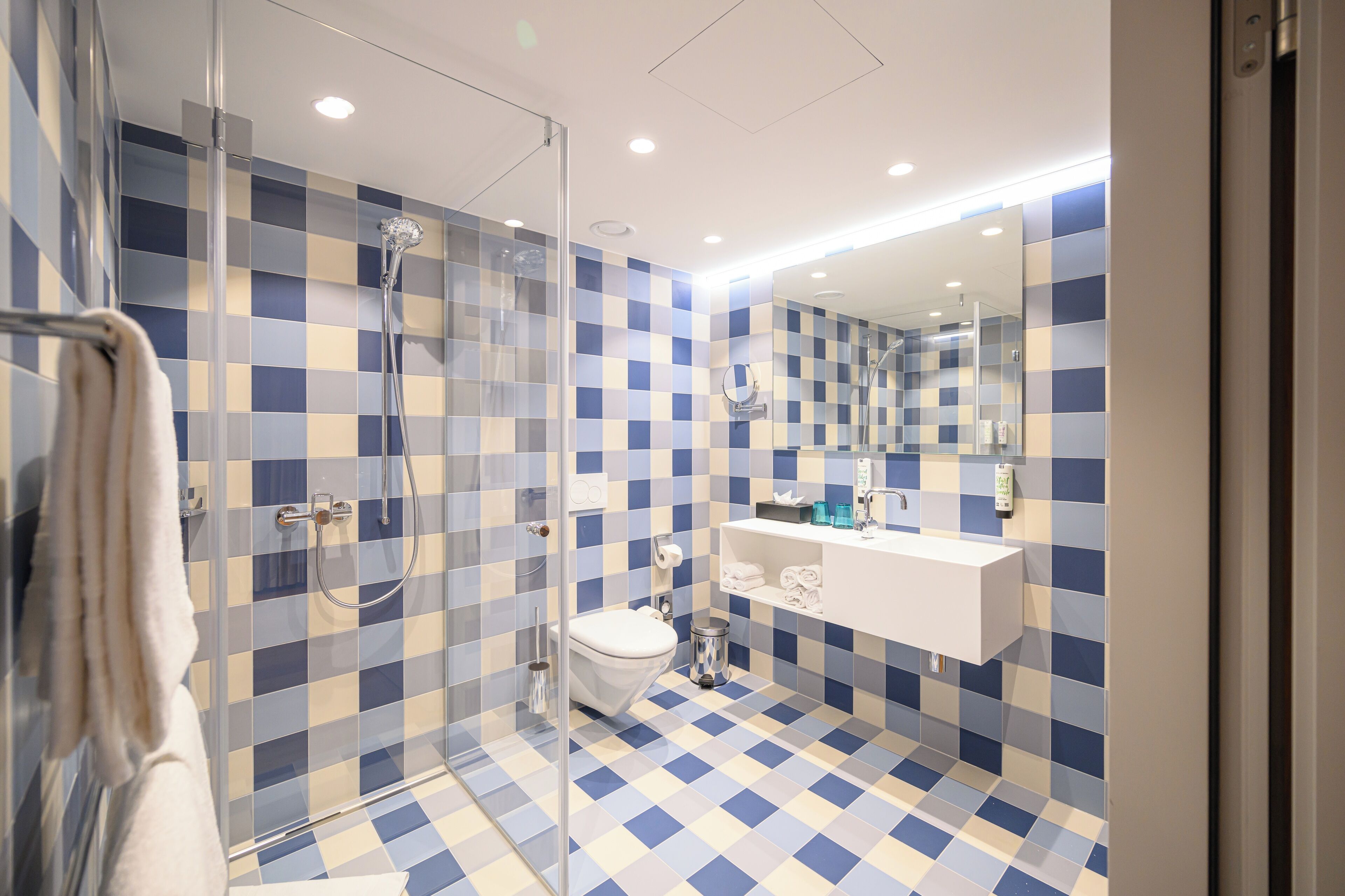 Une salle de bain moderne avec un carrelage à motifs à carreaux, une douche en verre, des toilettes blanches et un lavabo blanc avec un miroir au-dessus.
