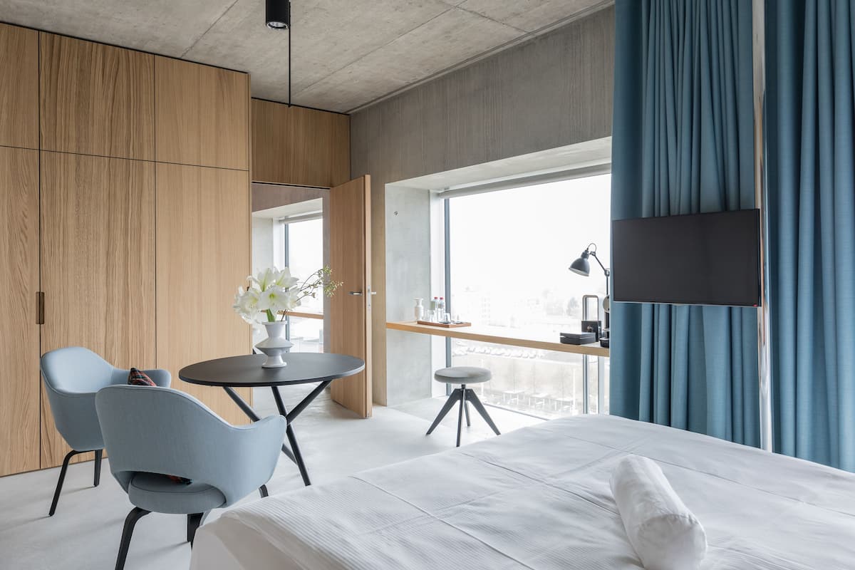 Une chambre d’hôtel moderne avec un grand lit, un bureau avec une chaise, une petite table ornée d’un vase de fleurs et une grande fenêtre avec des rideaux.