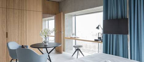 Een moderne hotelkamer met een groot bed, een bureau met stoel, een kleine tafel met een bloevas en een groot raam met gordijnen.