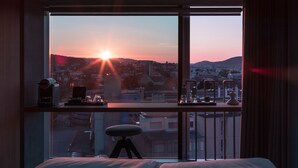 Une pièce dotée d’une grande fenêtre donnant sur un paysage urbain au coucher du soleil, d’un bureau avec une chaise et d’un lit.