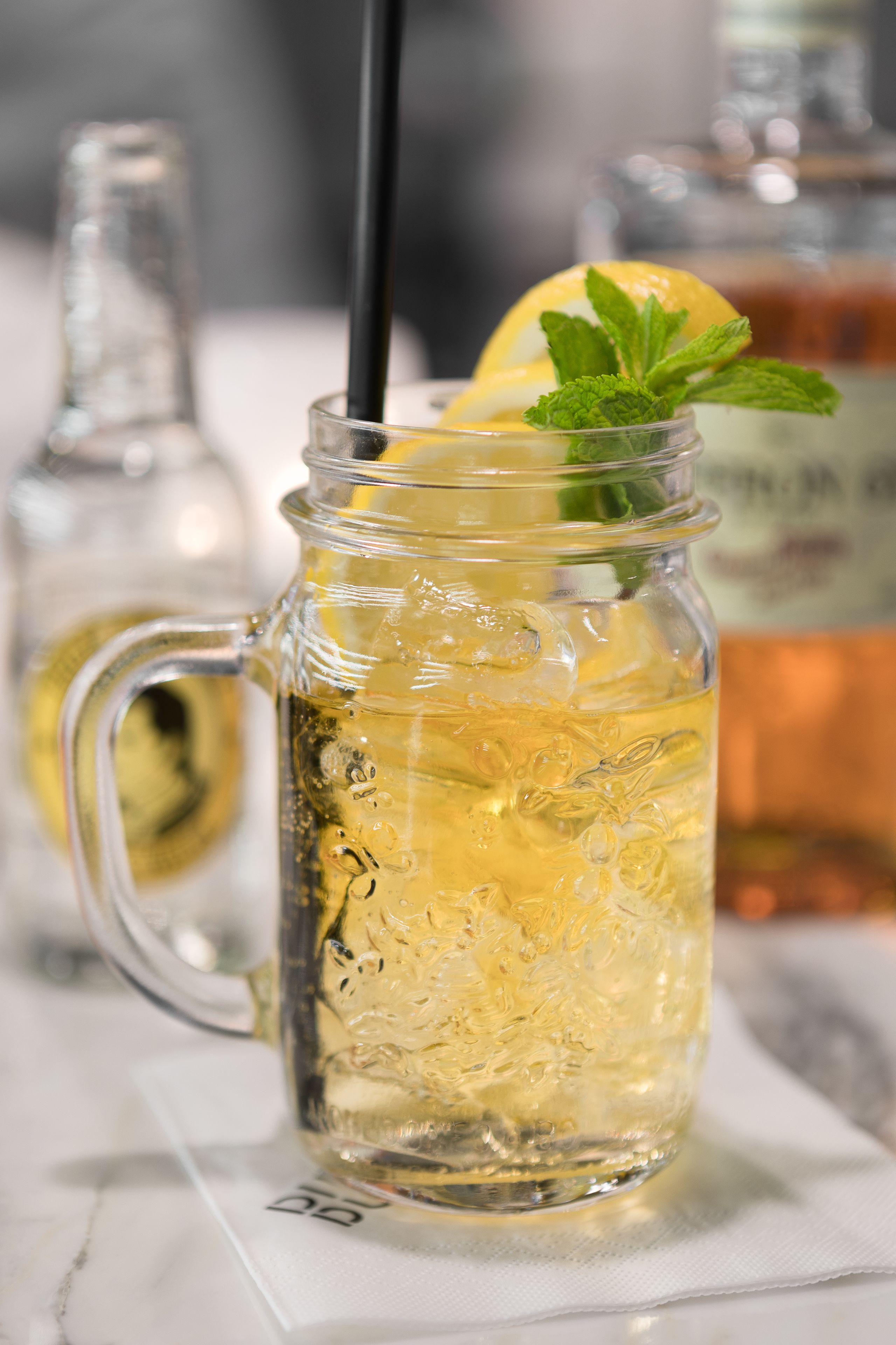 A clear glass mug with a handle, filled with a yellowish beverage, ice cubes, and garnished with a lemon slice and mint leaves.