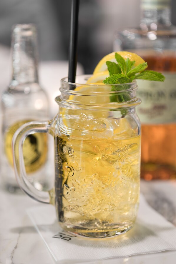 Una taza de vidrio transparente con asa, llena de una bebida amarillenta, cubitos de hielo y adornada con una rodaja de limón y hojas de menta.