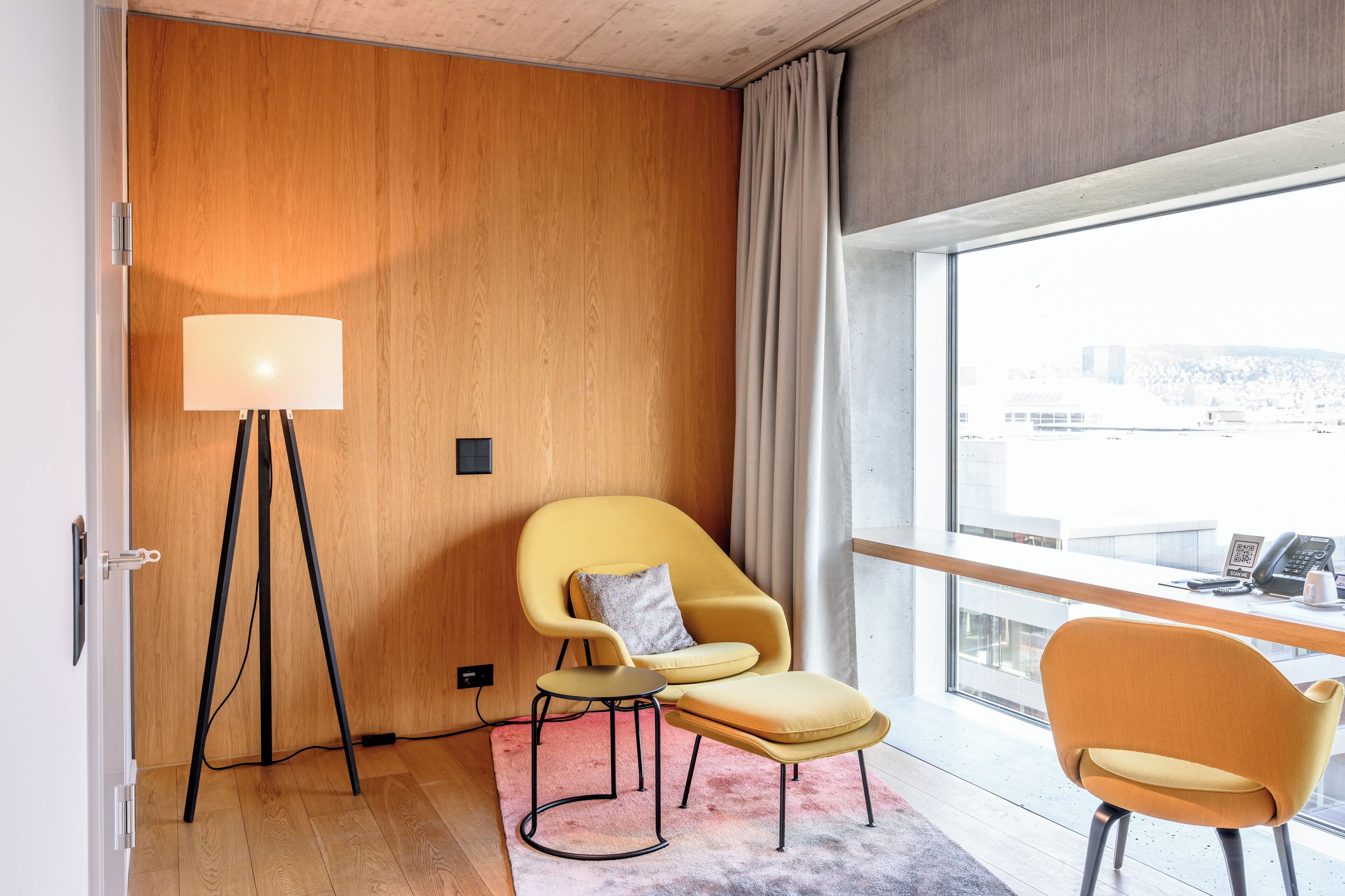 Ein modernes Hotelzimmer mit einer Holzwand, einem großen Fenster mit Blick auf die Stadt, einem gelben Sessel, einem kleinen runden Tisch und einer Stehlampe.
