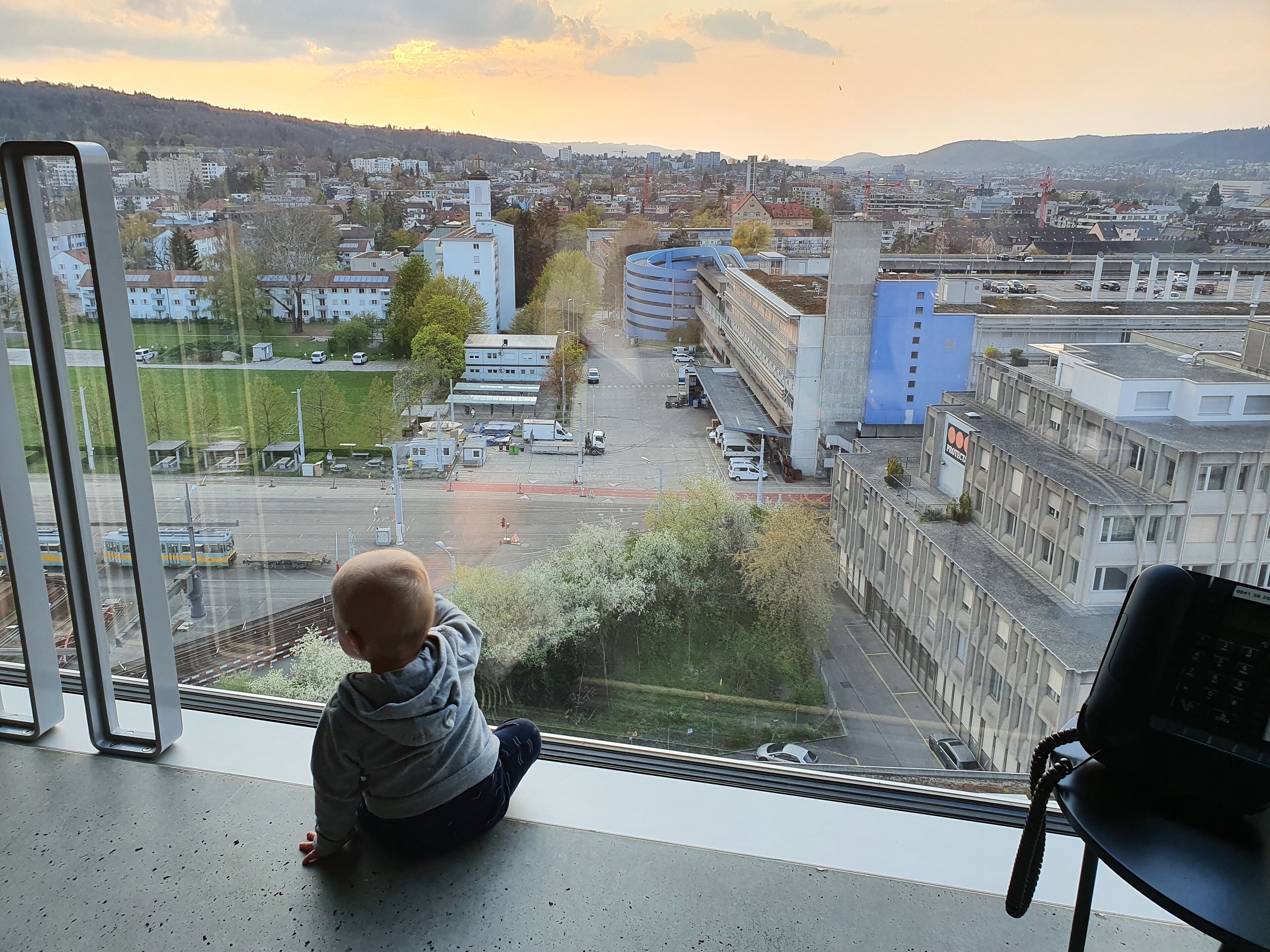 Un enfant est assis près d’une fenêtre donnant sur un paysage urbain, avec des immeubles, des routes et un tramway.