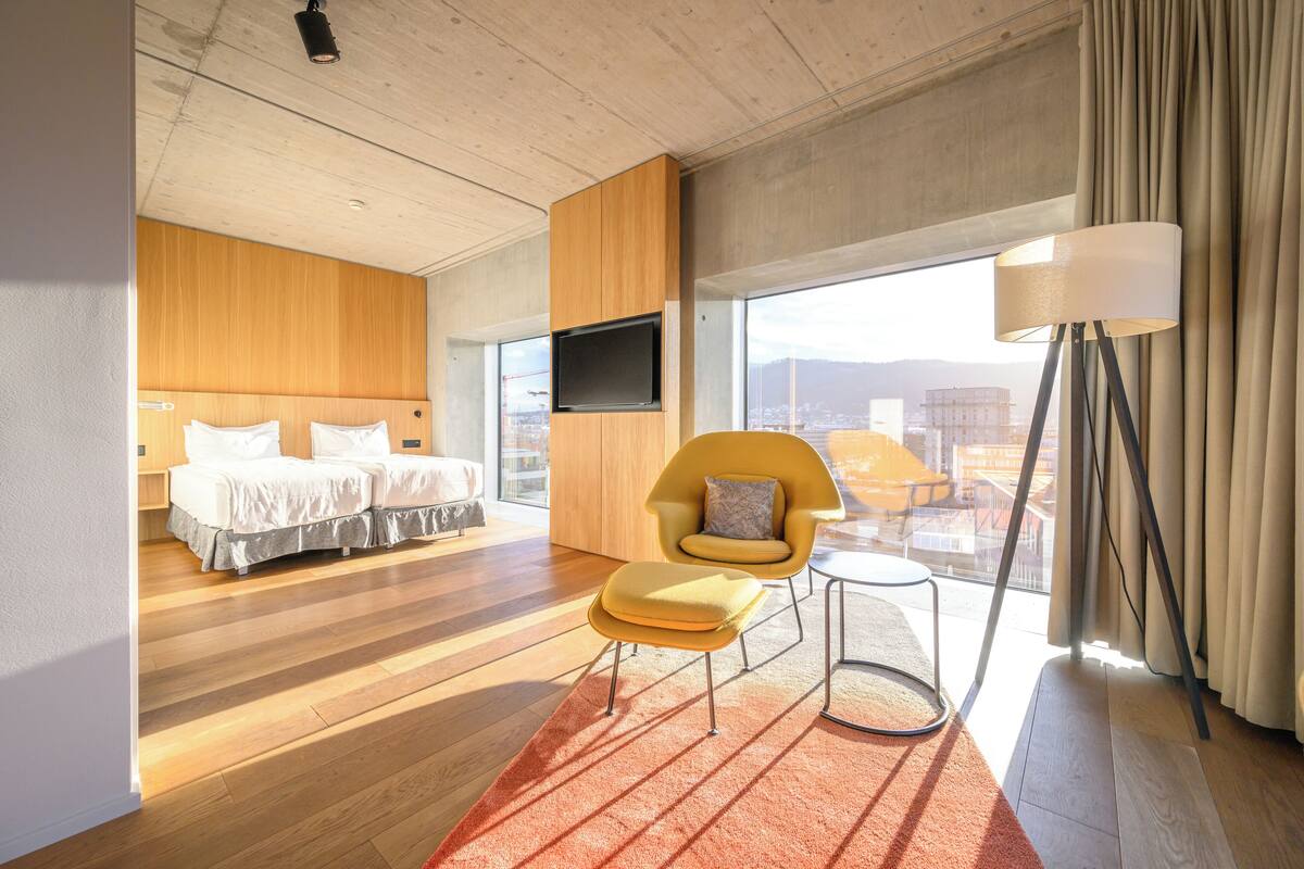 Une chambre d’hôtel moderne avec un plancher en bois, une grande fenêtre donnant sur un paysage urbain, un fauteuil jaune et un lit avec du linge de maison blanc.