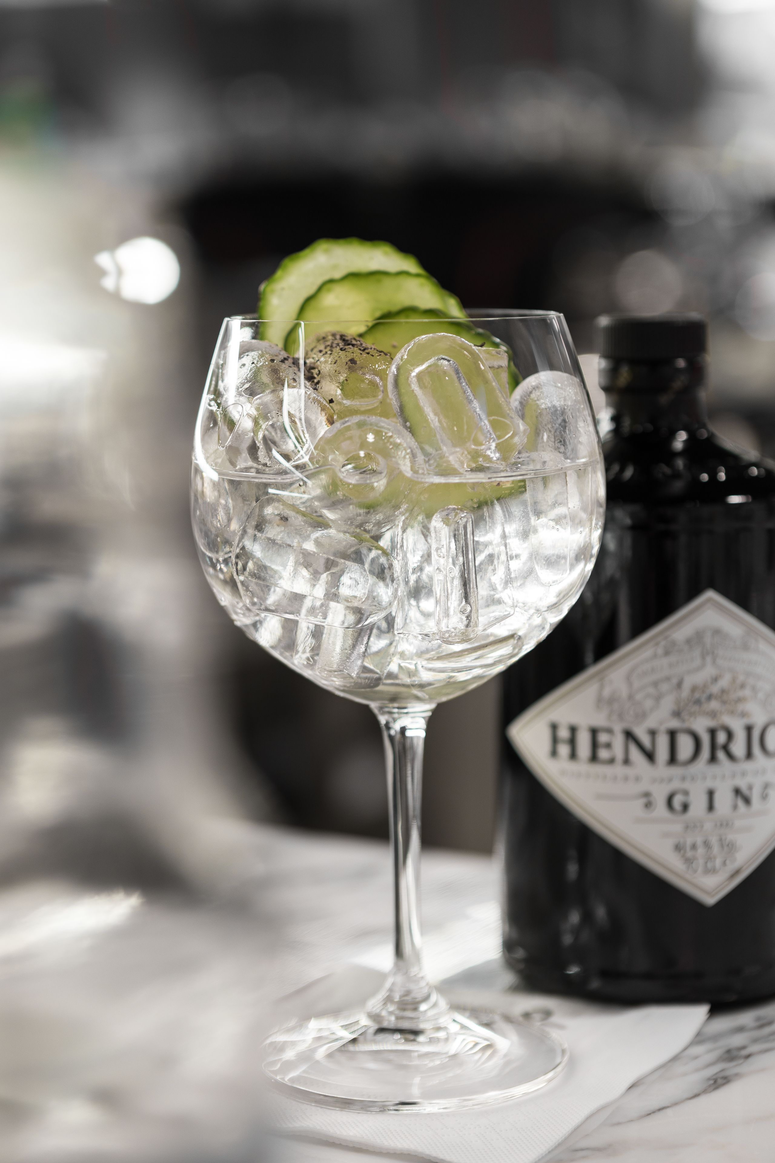 Un verre de gin avec des glaçons et des rondelles de concombre, une bouteille de gin Hendrick's et une surface en marbre.