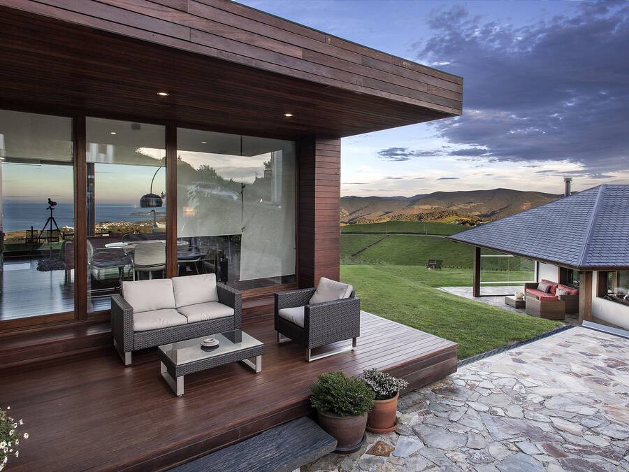 Un espacio al aire libre moderno con mobiliario de mimbre, un patio de piedra y una vista panorámica del océano y las colinas.