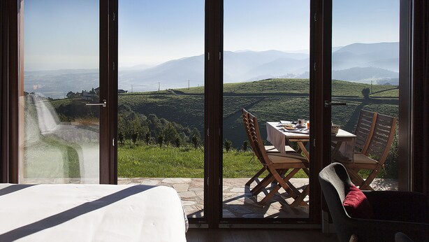 Una habitación con un gran ventanal corredera que da a un balcón con mesa y sillas, y vistas a un valle y montañas.