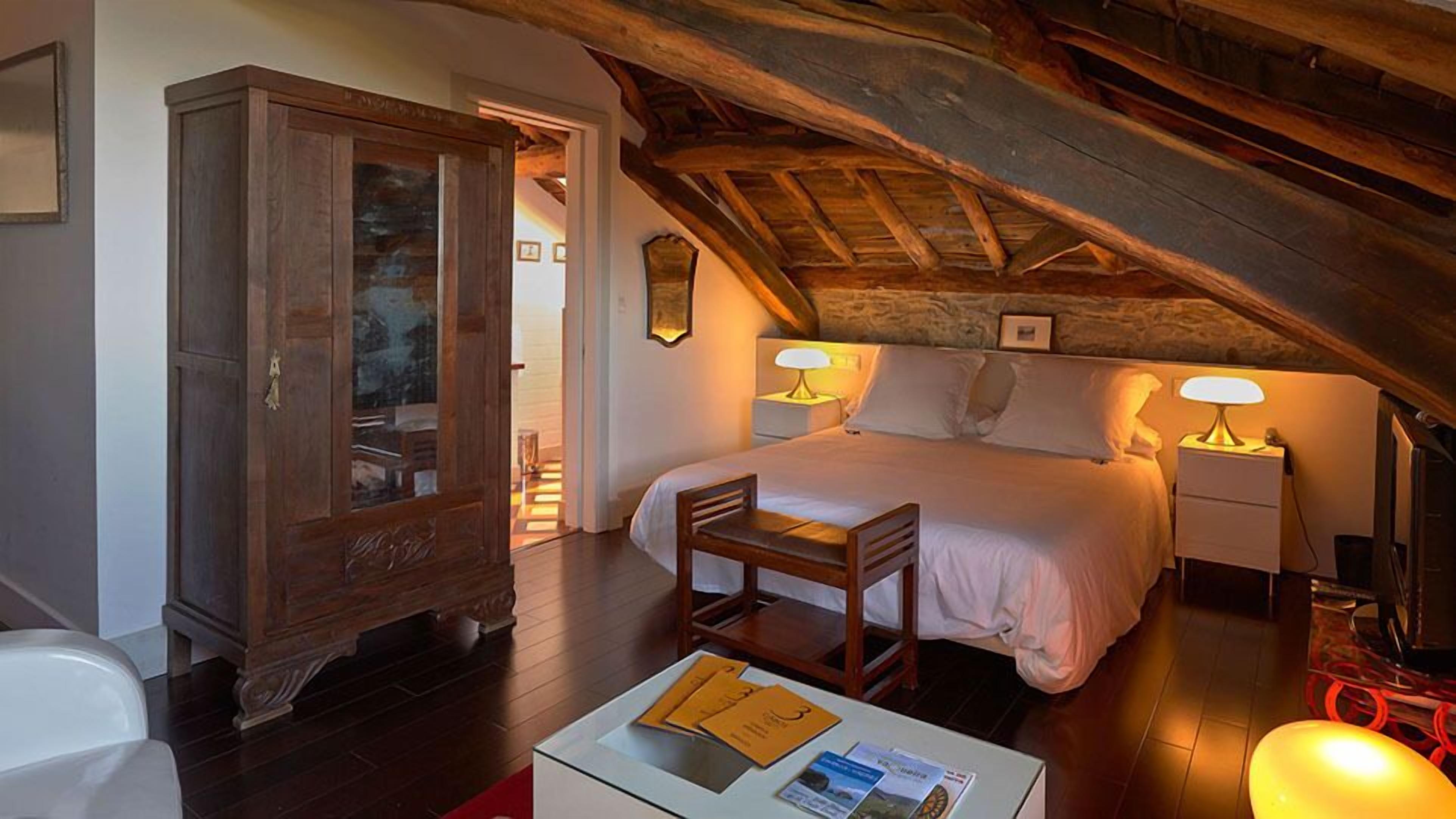 Ein Schlafzimmer mit Holzboden, einem Bett mit weißen Leinen, zwei Nachttischlampen, einem Holzstuhl und einem Schrank mit Glastüren.