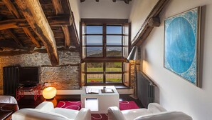 Habitación acogedora con pared de piedra, vigas de madera, un ventanal con vistas, un televisor de pantalla plana y una mesa con sillas.