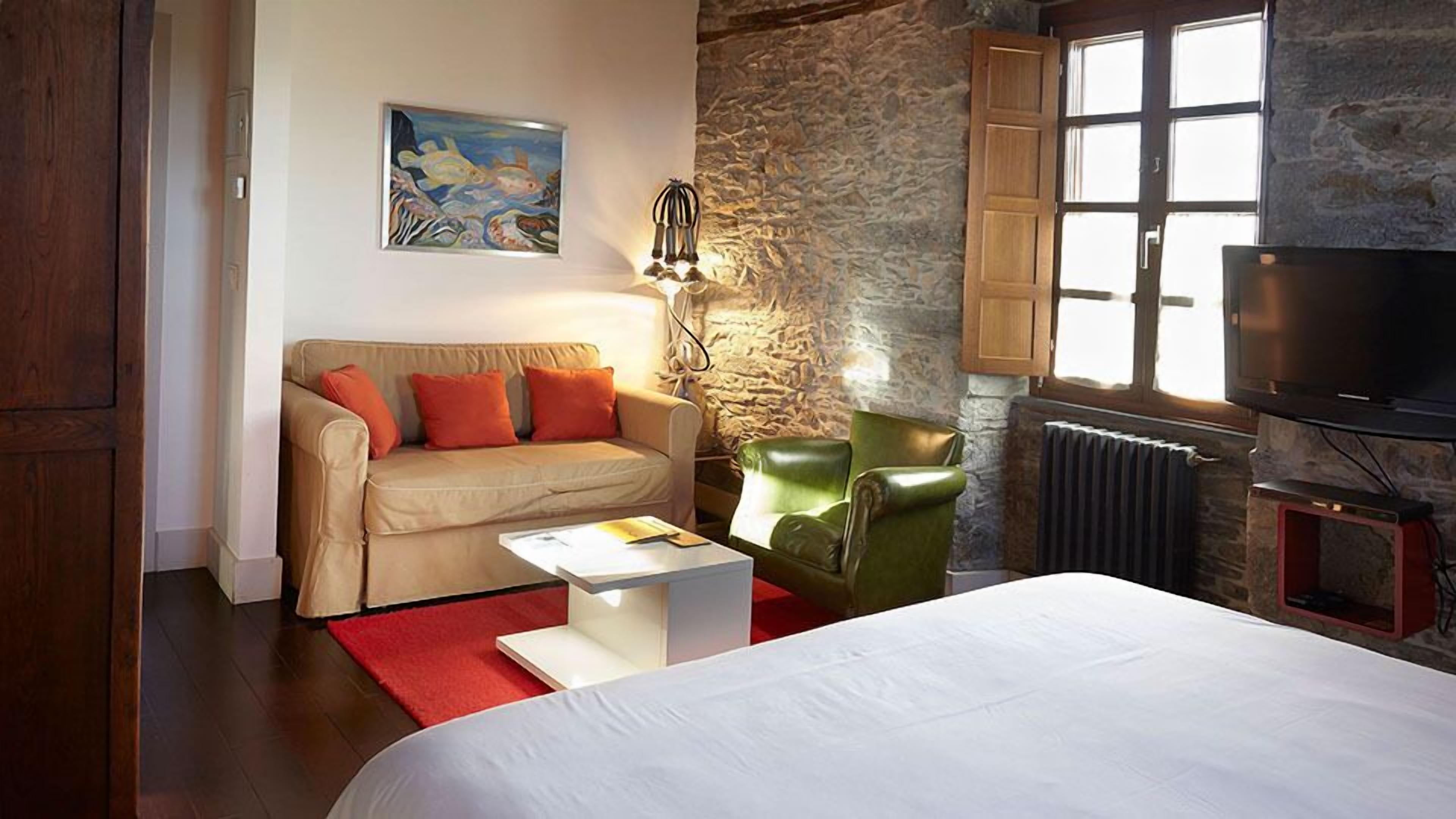 Habitación de hotel con cama, sofá, sillón verde, alfombra roja, un cuadro, pared de piedra y televisor.