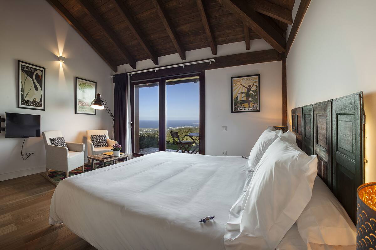 A bedroom with a large bed, a desk, a chair, and a TV. There are two windows with a view of the outdoors.