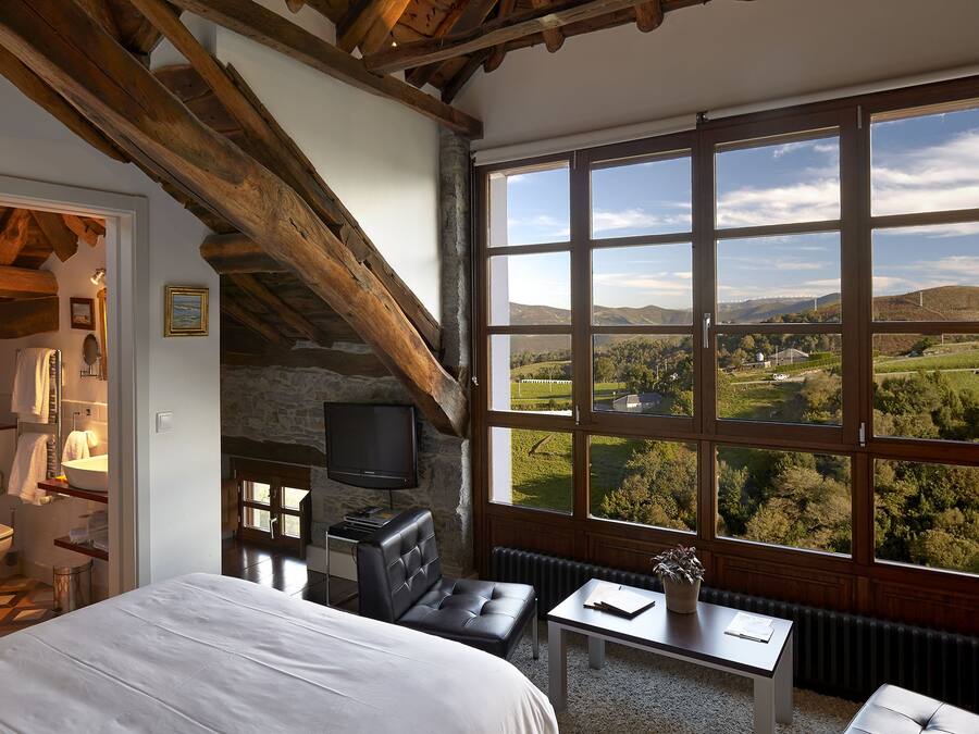 A bedroom with a large window, a bed, a small table, a chair, and a television.