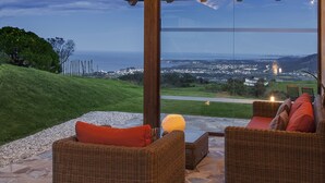 Un sofá de mimbre con cojines anaranjados y una lámpara de mesa, con vistas a un paisaje costero que incluye el mar y edificios lejanos.