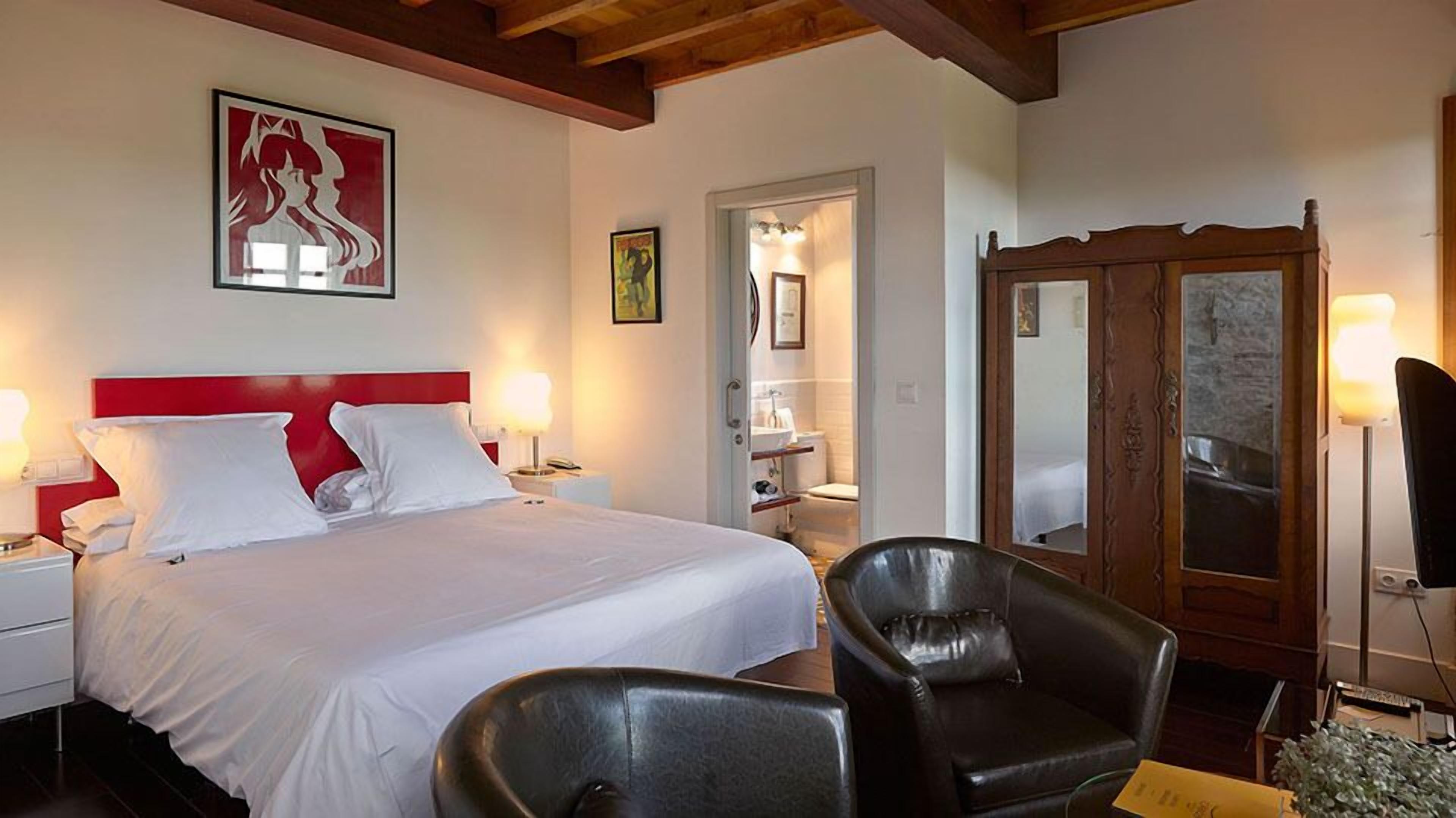 A hotel room with a large bed, a wooden wardrobe, a chair, and a bathroom visible through an open door.