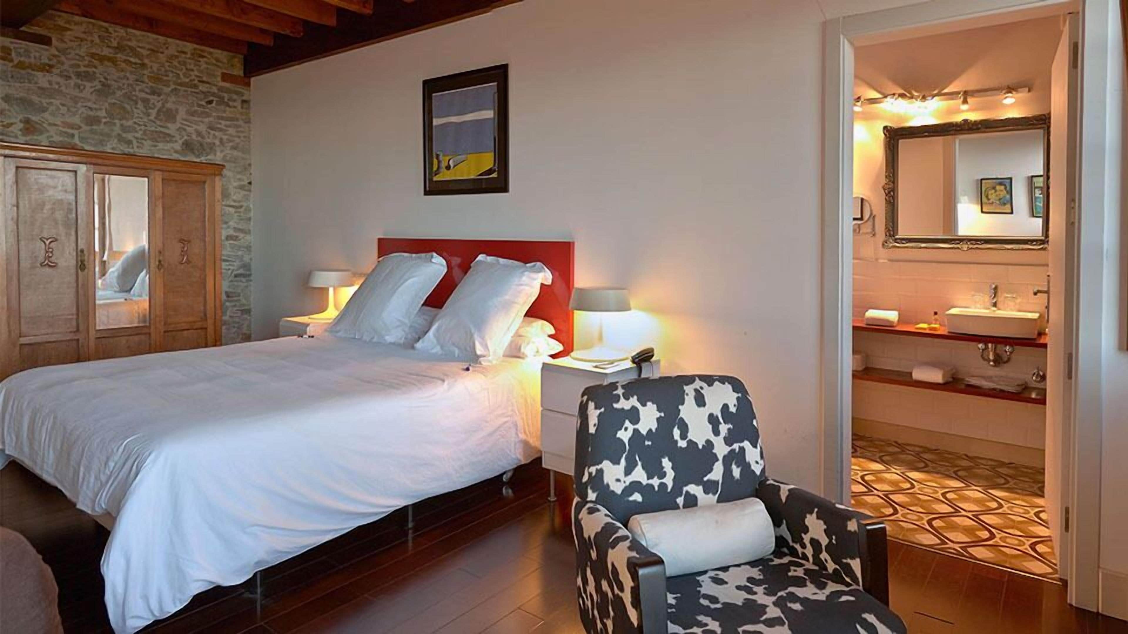 Habitació d'hotel amb un llit, una cadira, una taula de nit, una làmpada, un quadre i un bany amb un lavabo i un mirall.