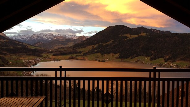 Vista desde un balcón con baranda de madera, sobre un valle con un lago, montañas y un pequeño poblado.