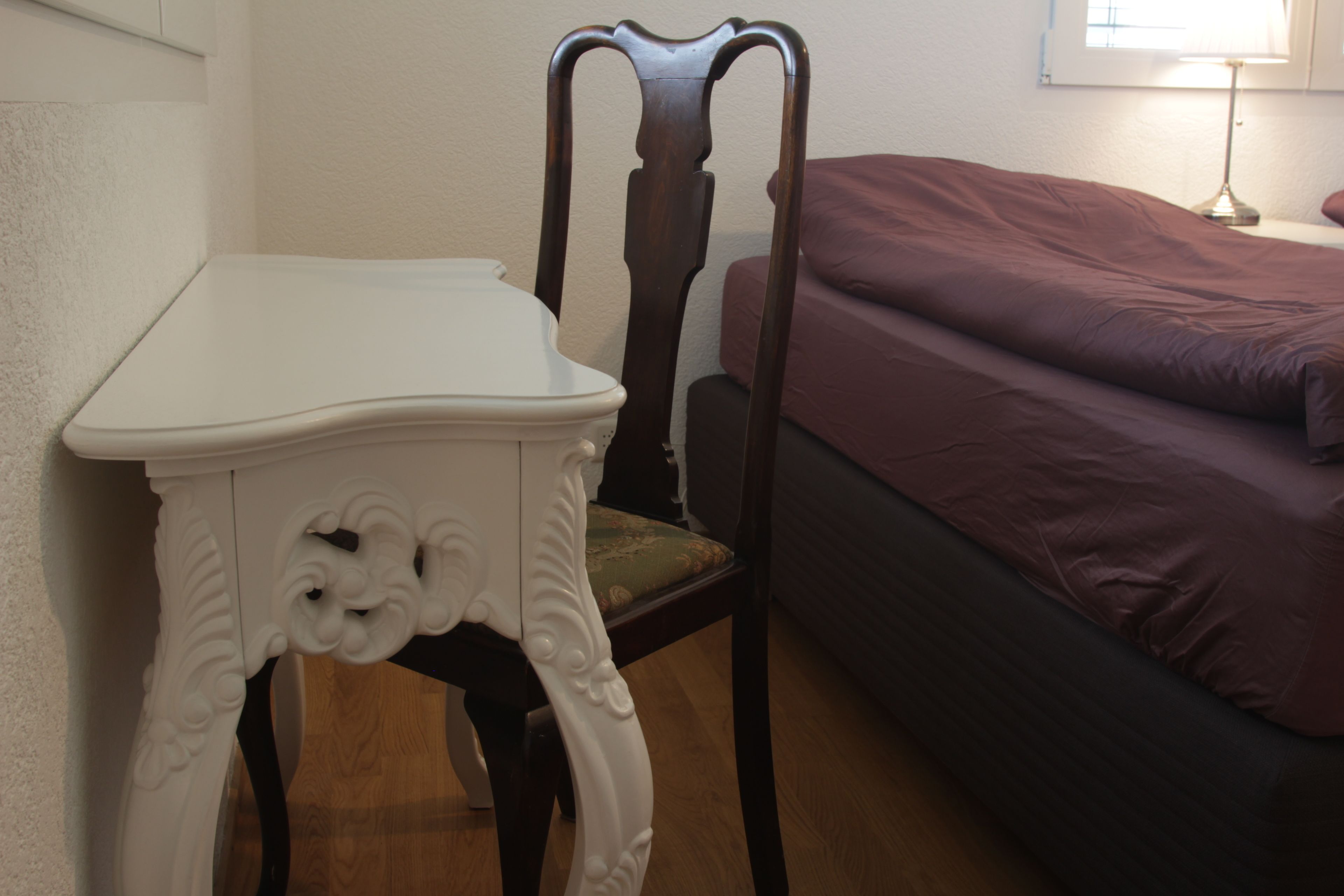 A room with a white table, a wooden chair, and a bed with a purple bedspread.
