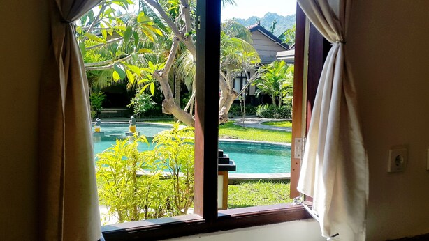 A view through a window with curtains, revealing a swimming pool, greenery, and a building in the background.