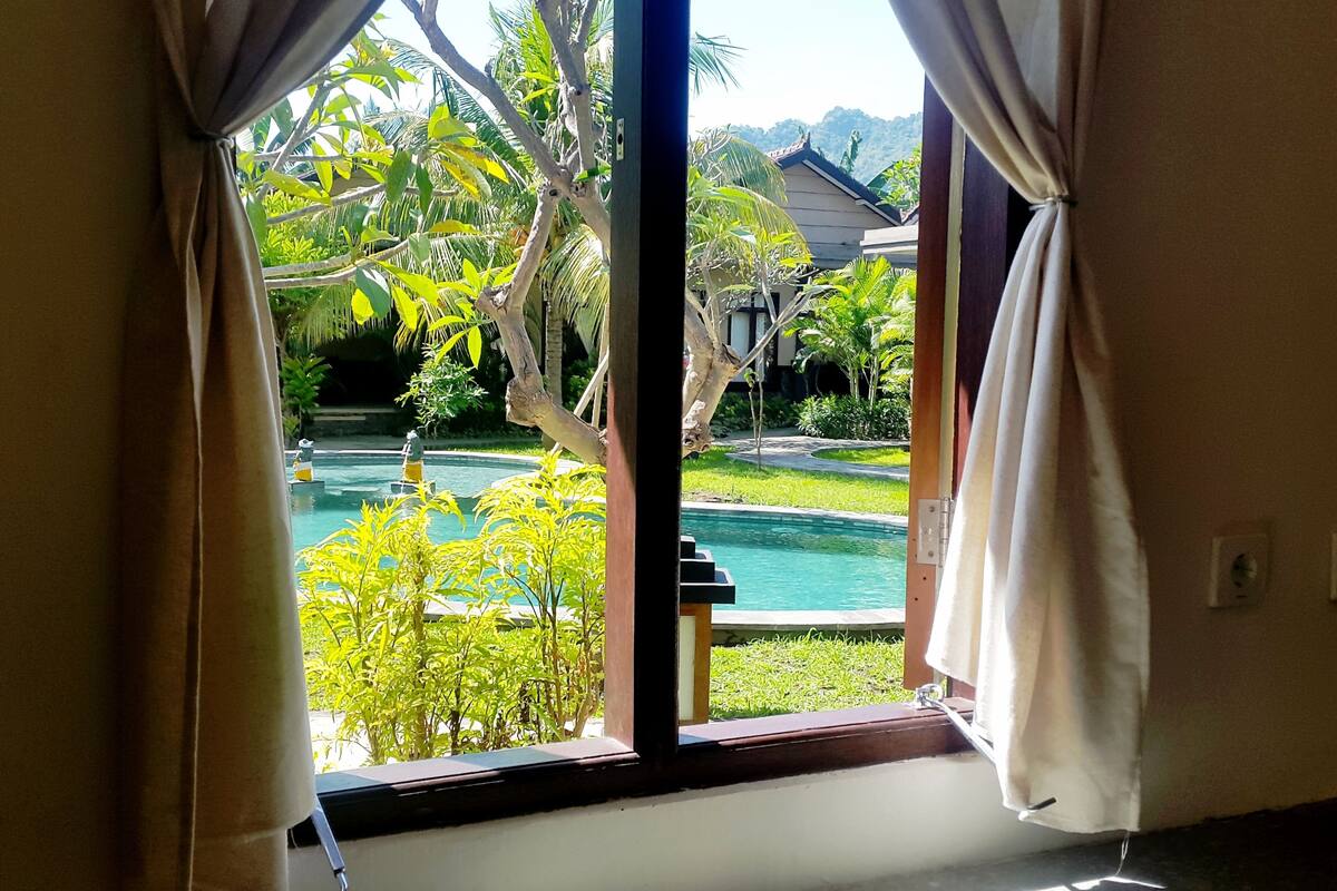 A view through a window with curtains, revealing a swimming pool, greenery, and a building in the background.