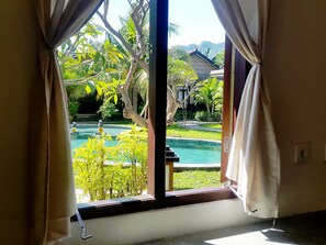 A view through a window with curtains, revealing a swimming pool, greenery, and a building in the background. - Doubleyou Home Stay Pemuteran (Pemuteran)