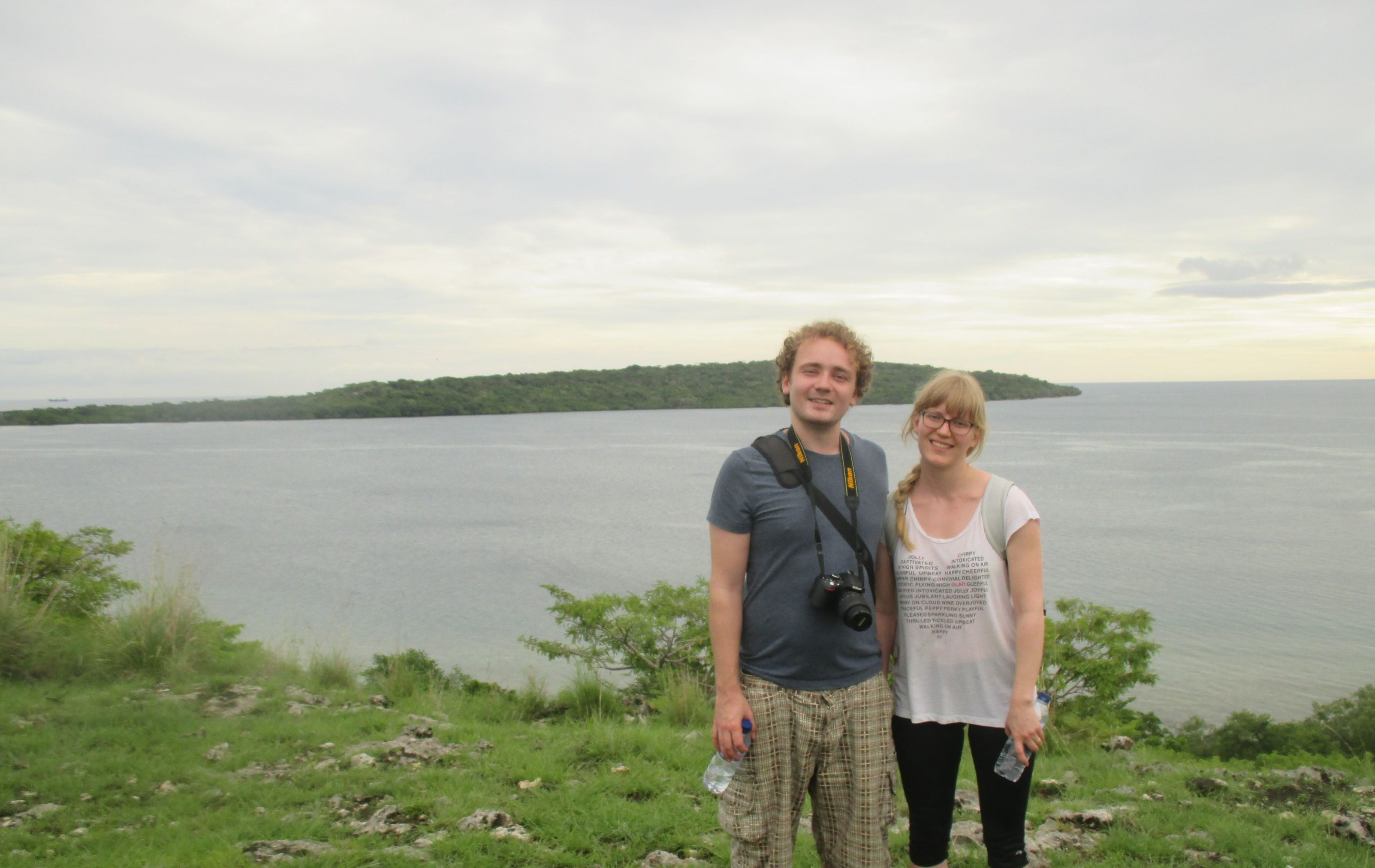 Two people standing on a grassy area with a lake and a distant island in the background.