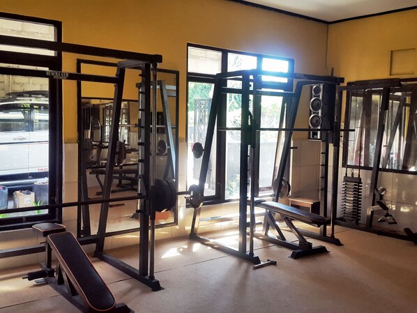 A gym with various weightlifting equipment, benches, and a window with a view of the outside. - Doubleyou Home Stay Pemuteran (Pemuteran)