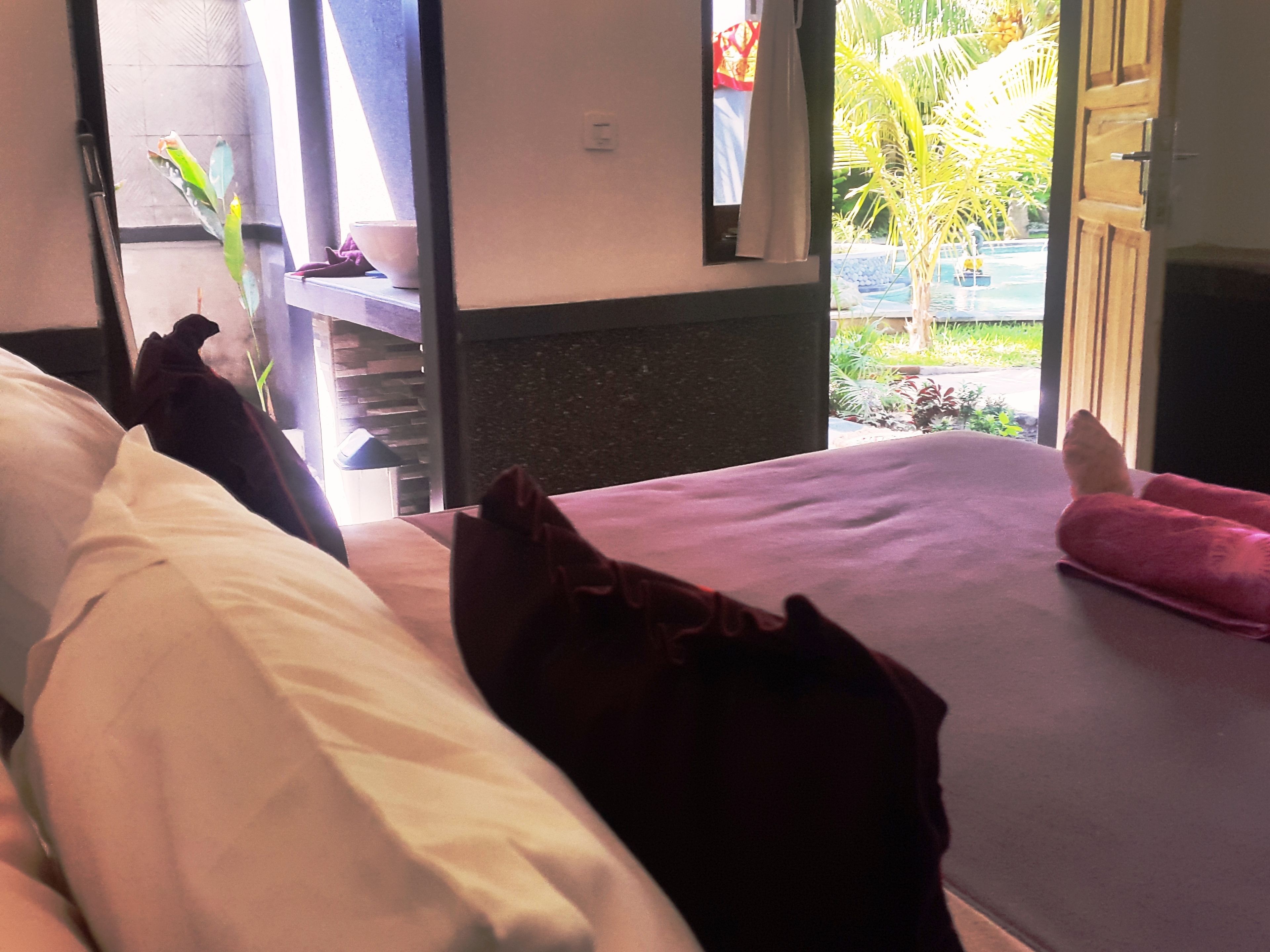 A hotel room with a bed, pillows, and a view of a pool and greenery outside.