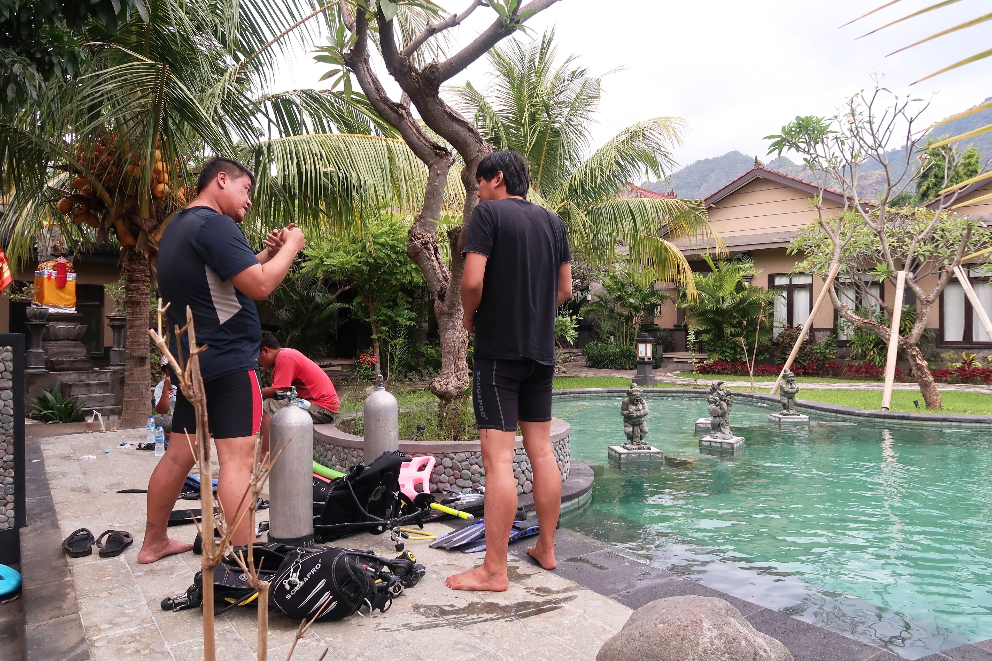 Two people near a pool with scuba diving gear and a statue in the water.