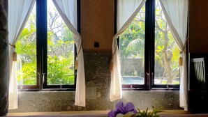 A room with two windows, each with white curtains, a stone wall, and a view of greenery and a pool outside.