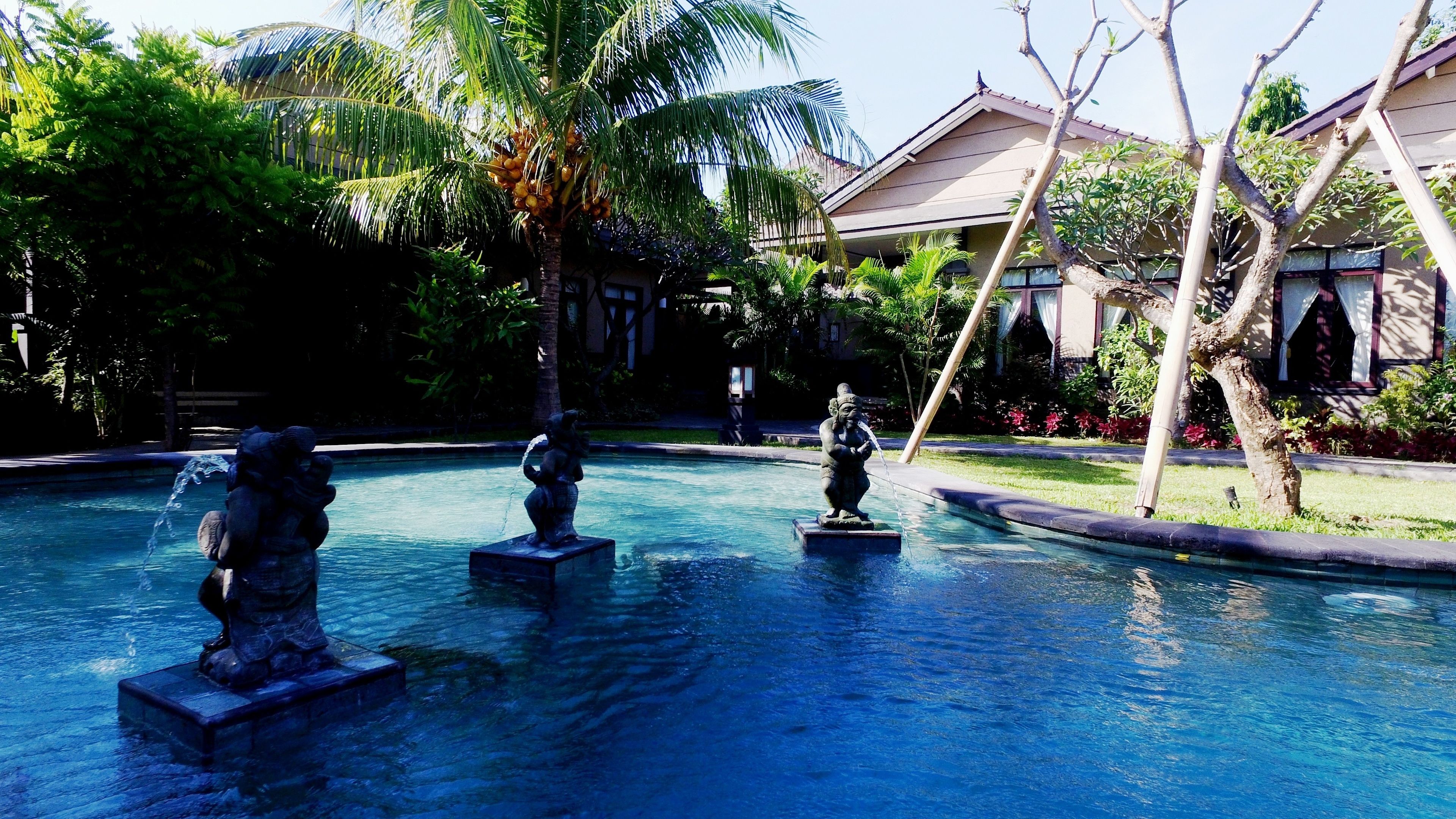A pool with statues, surrounded by palm trees and a building with a sloped roof.