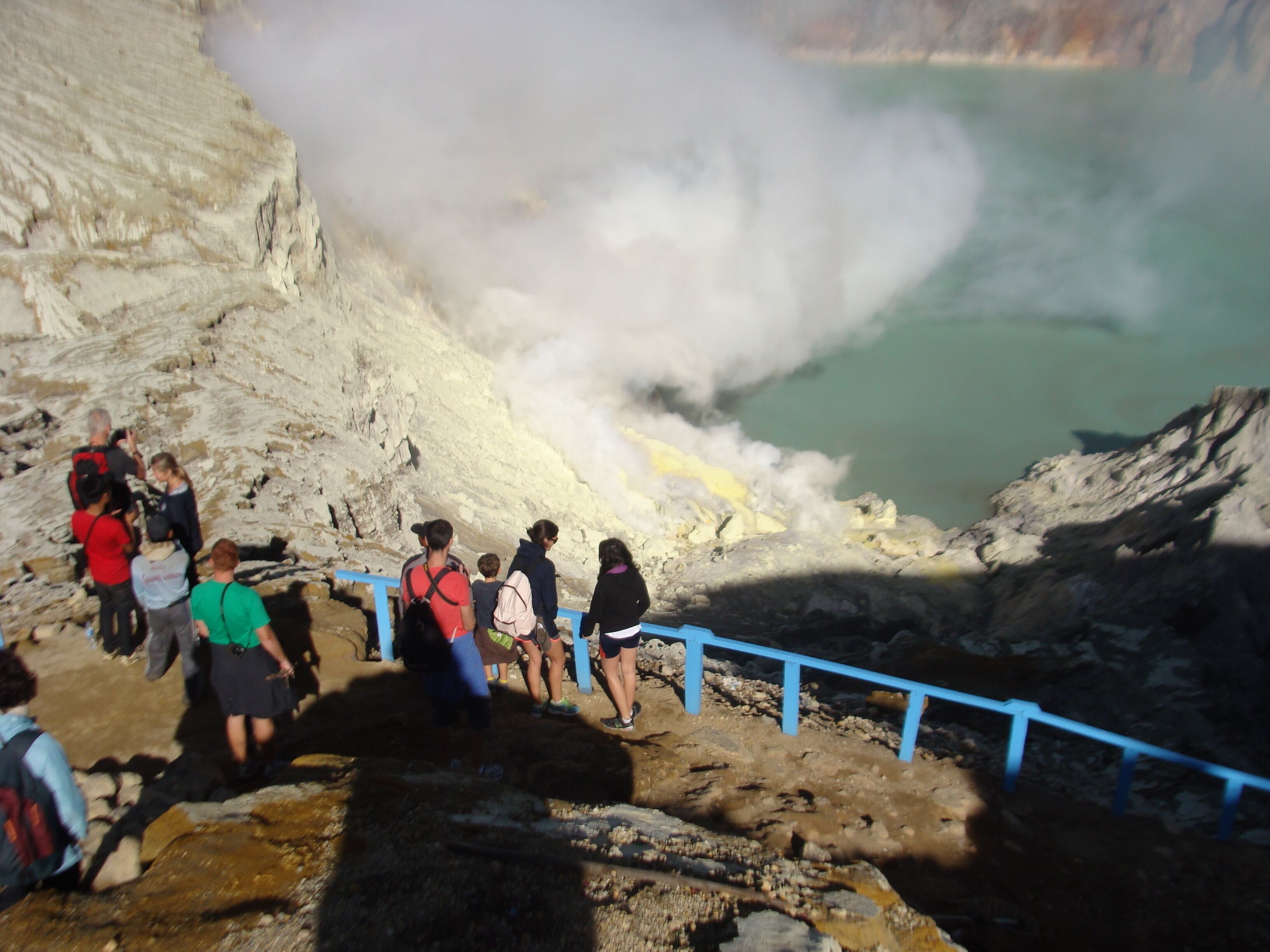 A group of people observing a geothermal feature with steam rising from the ground.