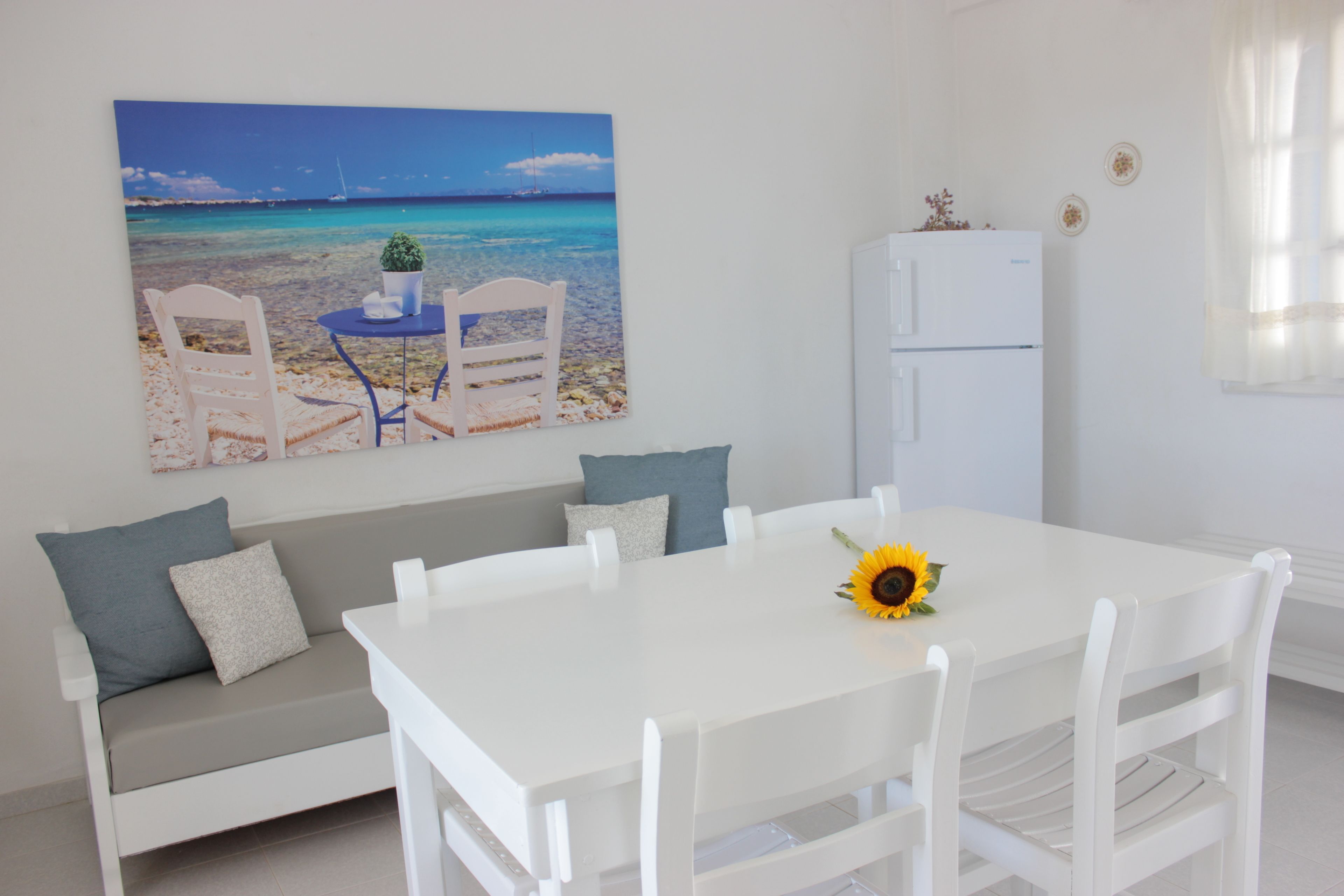 A dining area with a white table and chairs, a white refrigerator, a sofa with cushions, and a beach scene painting on the wall.