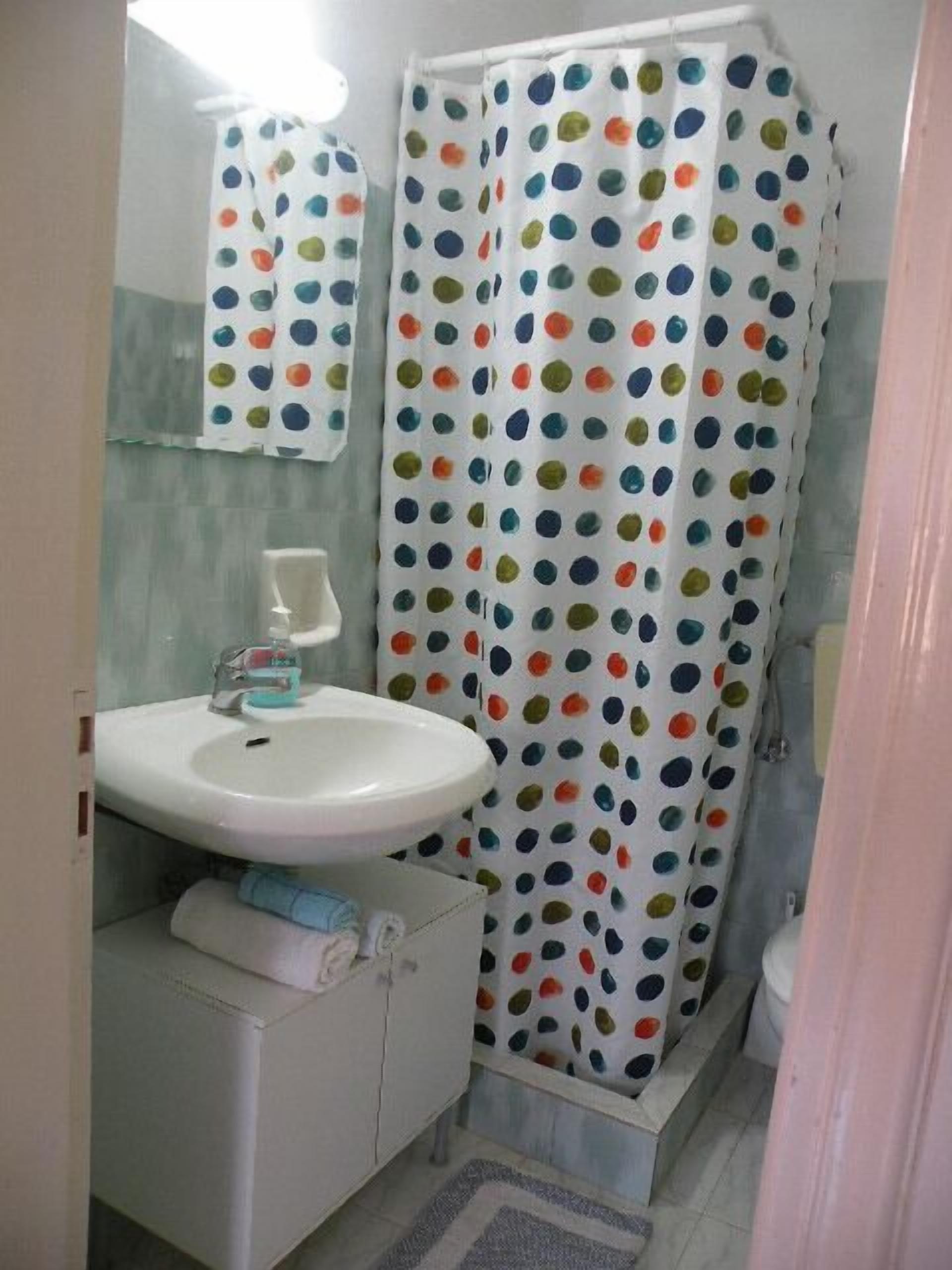 Un baño con lavamanos blanco, un espejo, una cortina de ducha con lunares de colores y un perchero.