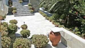 A white building with a blue door, a staircase leading to the entrance, and a garden with potted plants and a large terracotta pot.