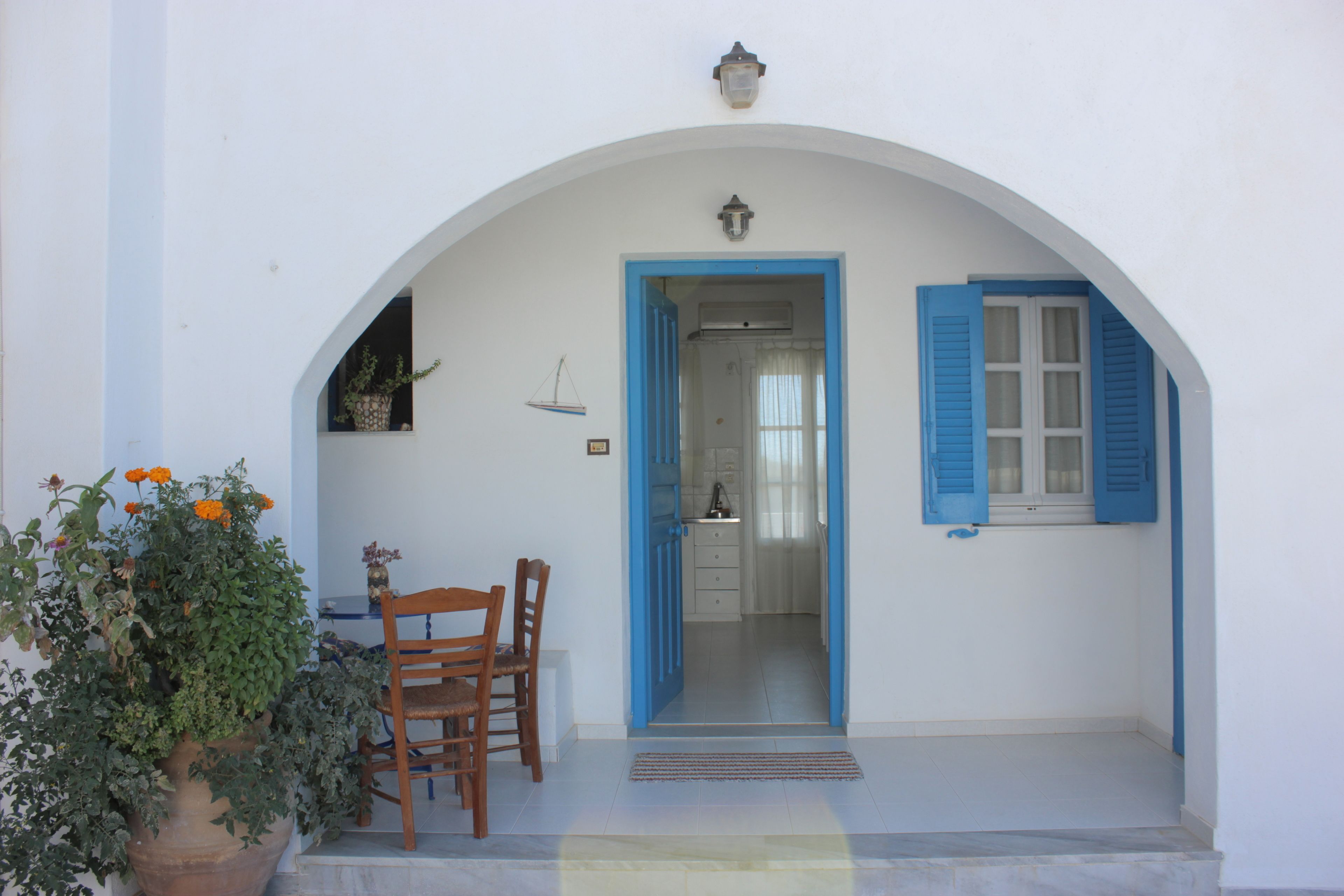A white building with a blue door and window shutters, a wooden table and chair, and a potted plant with orange flowers.