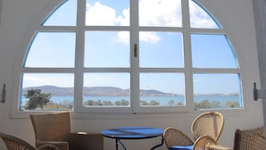 A room with a large arched window offering a view of the sea and distant hills. There are wicker chairs and a small round table.