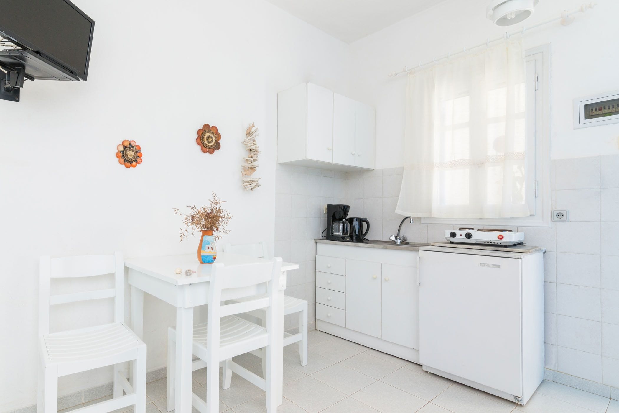 Uma cozinha pequena e clara, com geladeira branca, cafeteira e mesa branca com duas cadeiras.