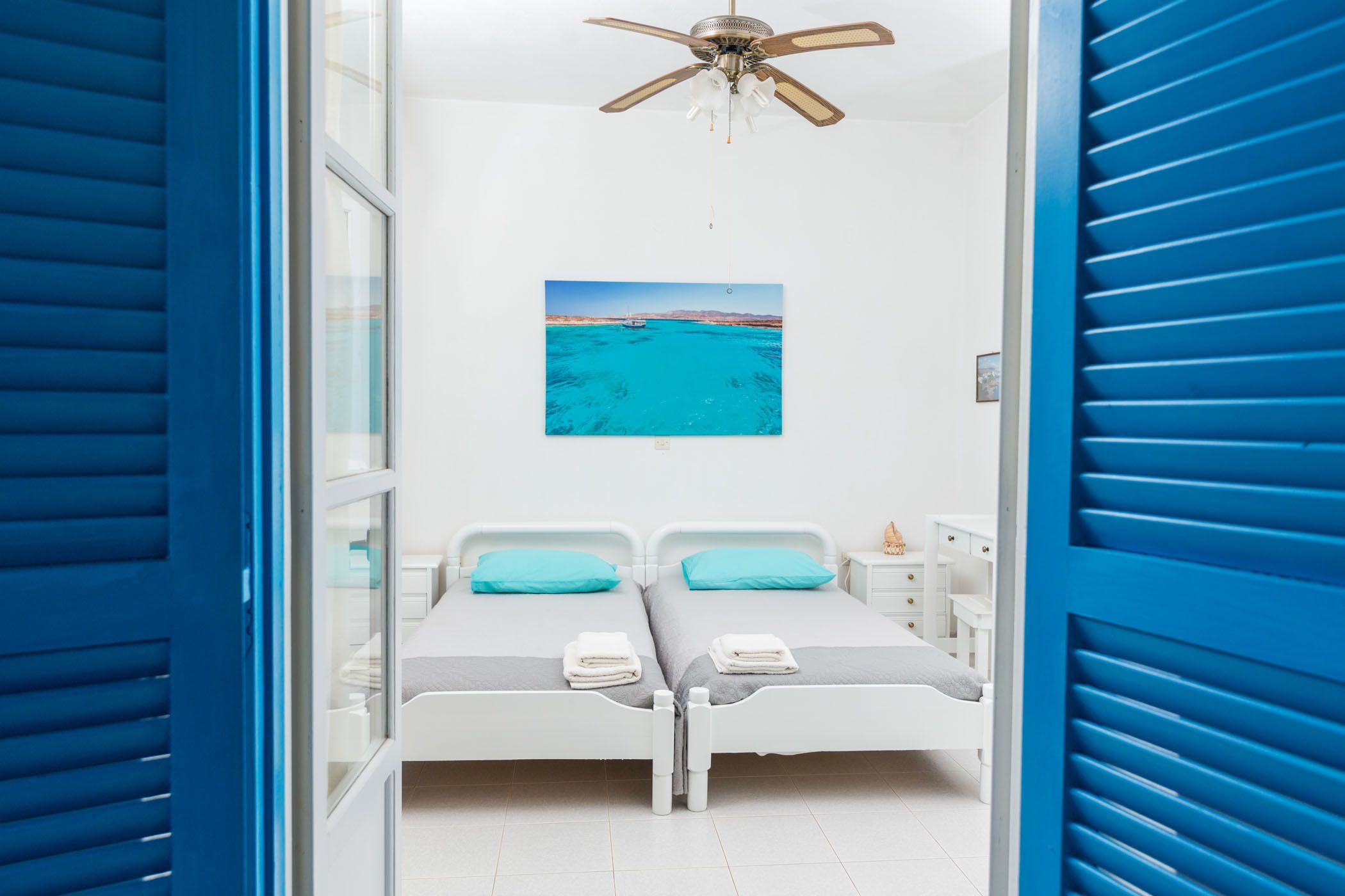 A room with two beds, a ceiling fan, a painting of a beach, and a window with blinds.