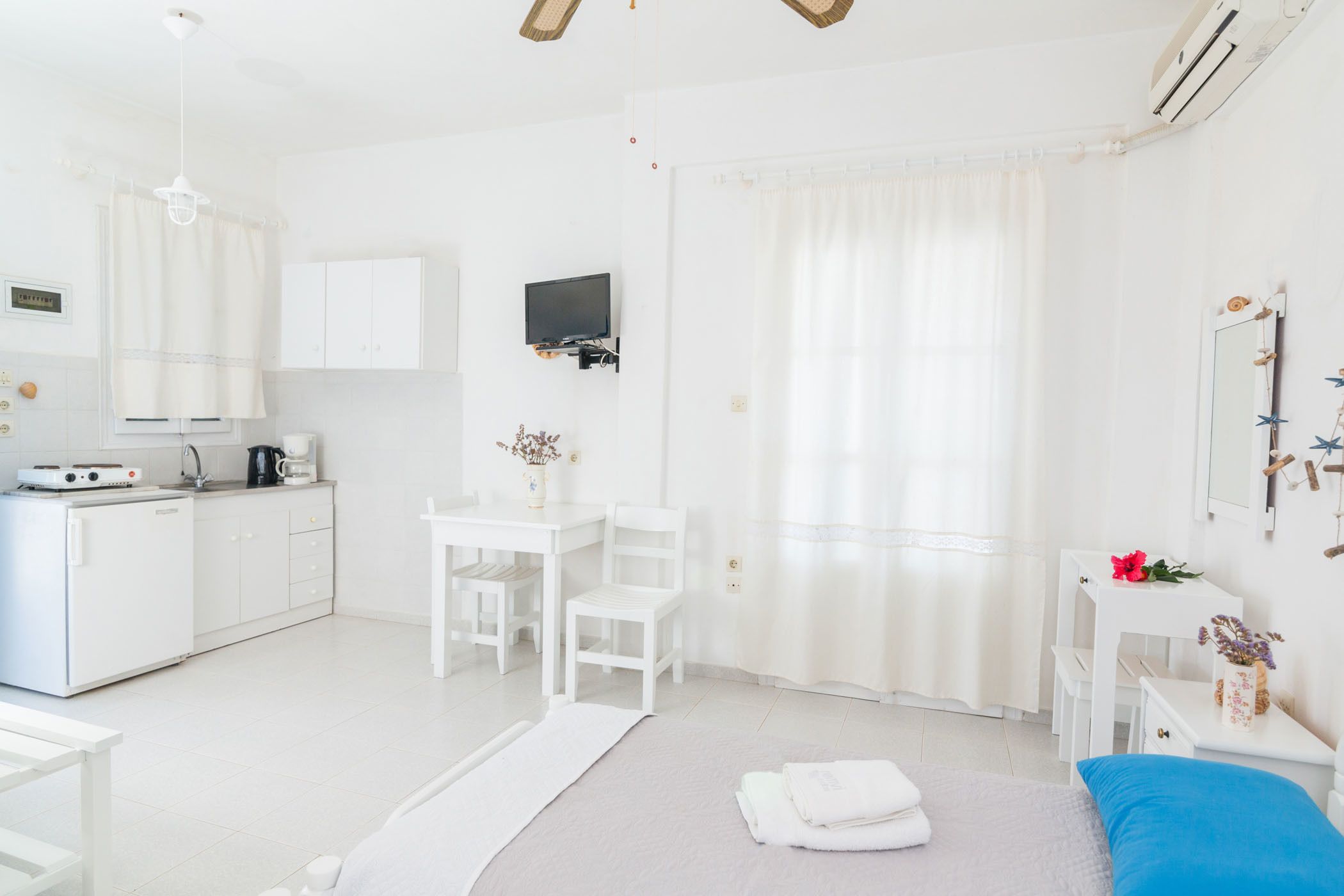 A white room with a bed, a desk, a chair, a TV, and a kitchenette.